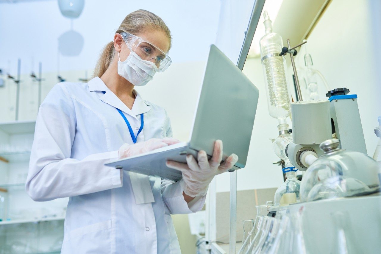 Serious confident young female scientist in protective mask and safety goggles using laptop to examine file with experiment result while working by medical equipment